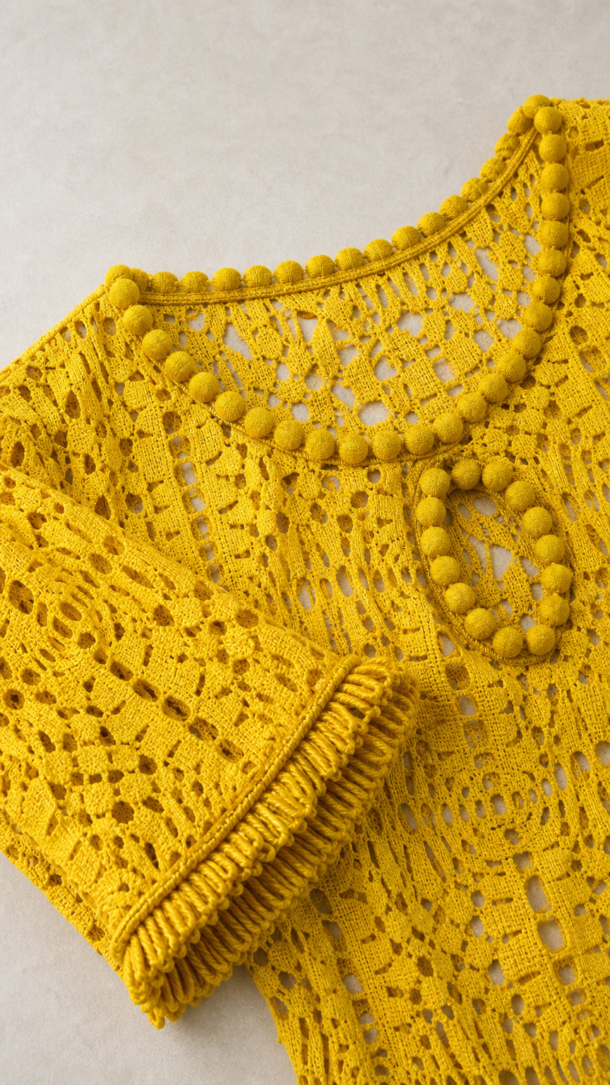 Close-up detail of yellow crochet knit pattern, pom-pom neckline, and sleeve cuff