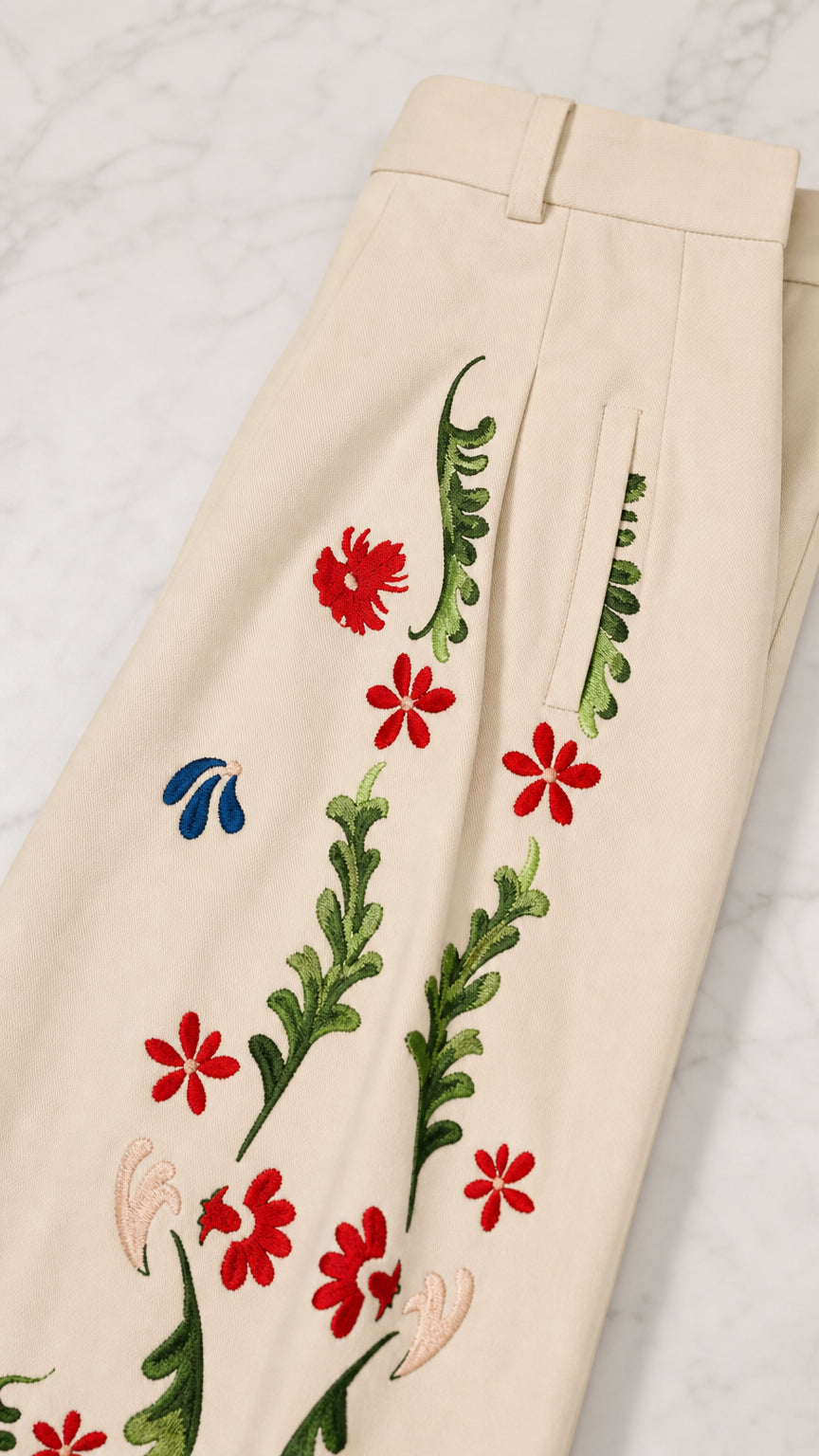 Close-up detail of high waistband, tailored pleats, pocket, and colorful floral embroidery on cream trousers